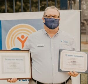 A man with a mask holding two certificates in front of a fence.