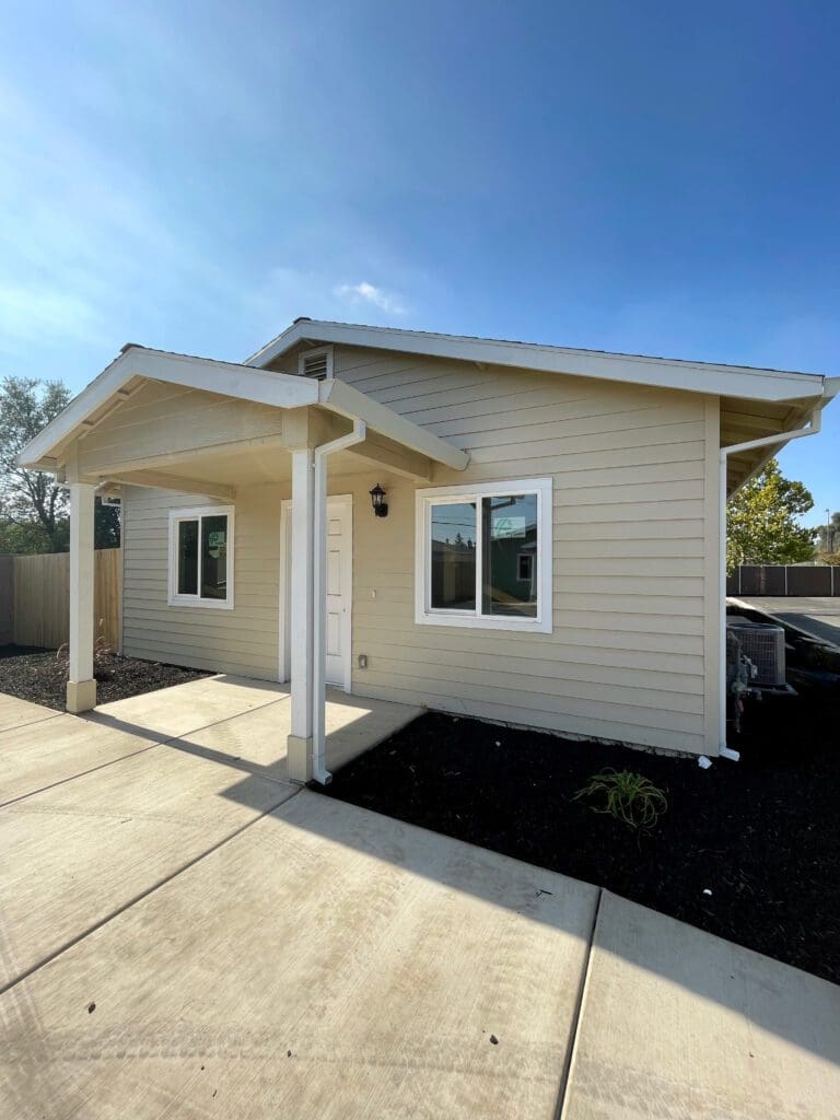 Exterior view of a small, modern single-story house with beige siding, white trim, and a covered porch, situated under a clear sky.